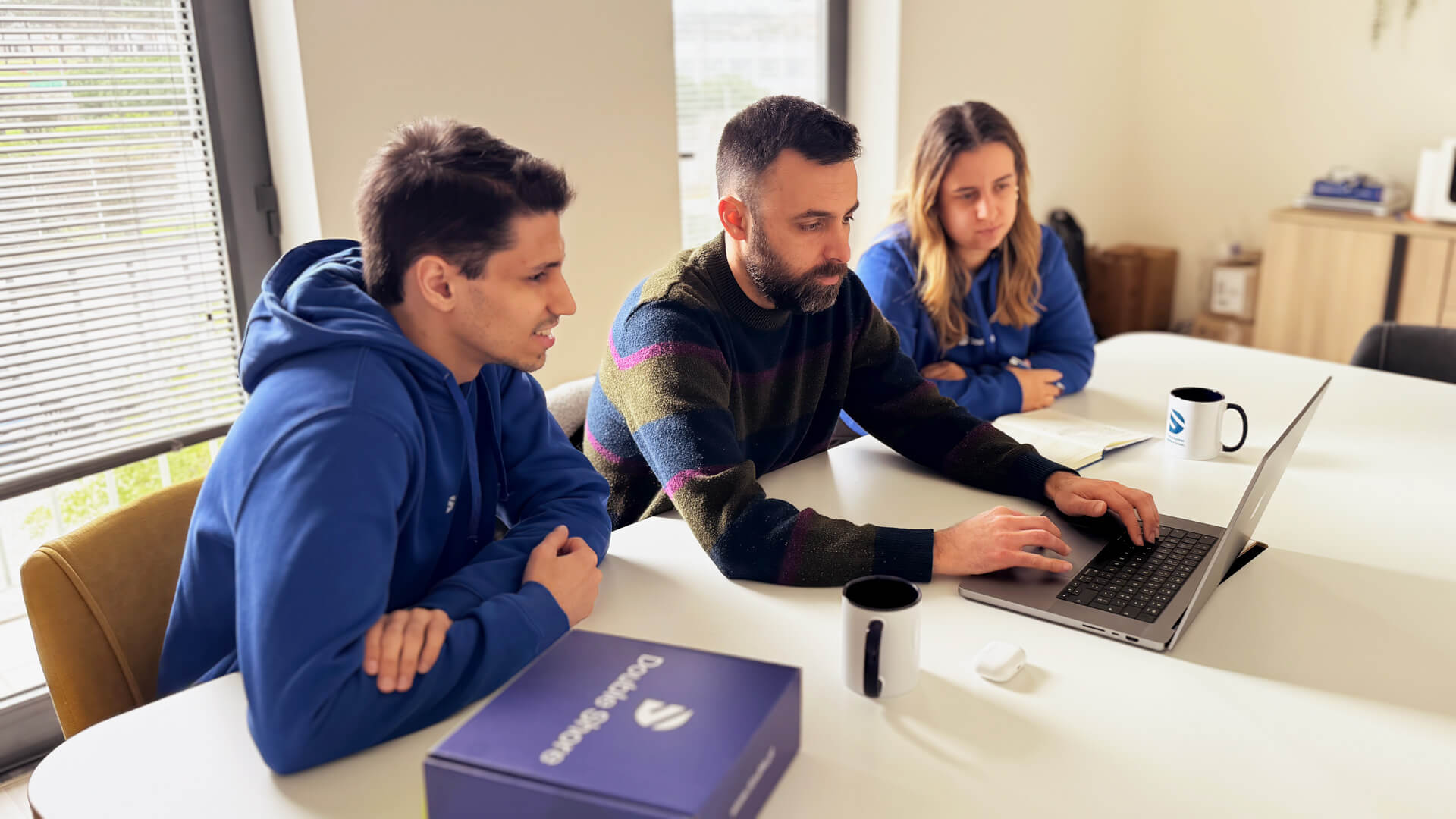 Três colegas reunidos à mesa analisam dados num portátil, colaborando num projeto em ambiente de escritório luminoso.